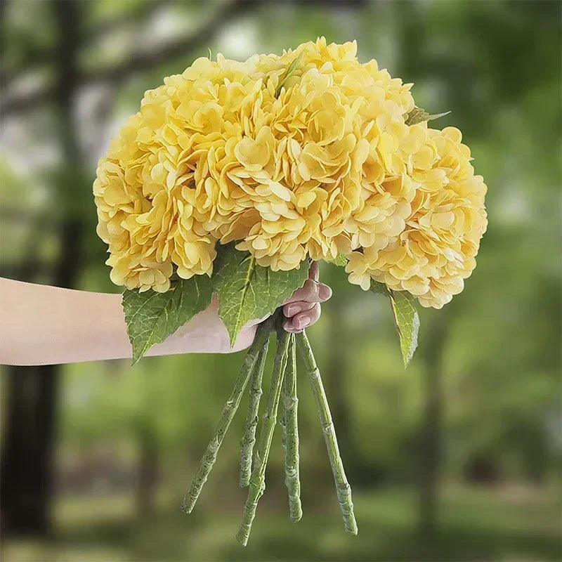 A hand holds a large cluster of yellow artificial hydrangea flowers with green stems against a blurred green background.
