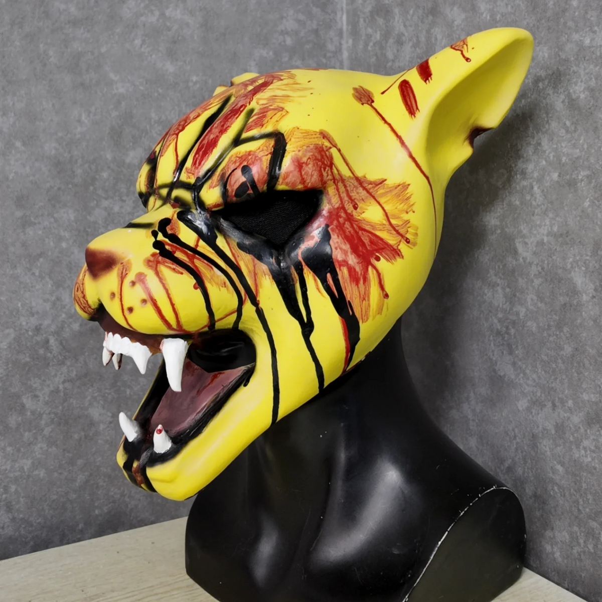 A yellow latex mask resembling a tiger's face with blood splatters and teeth, displayed on a black bust.