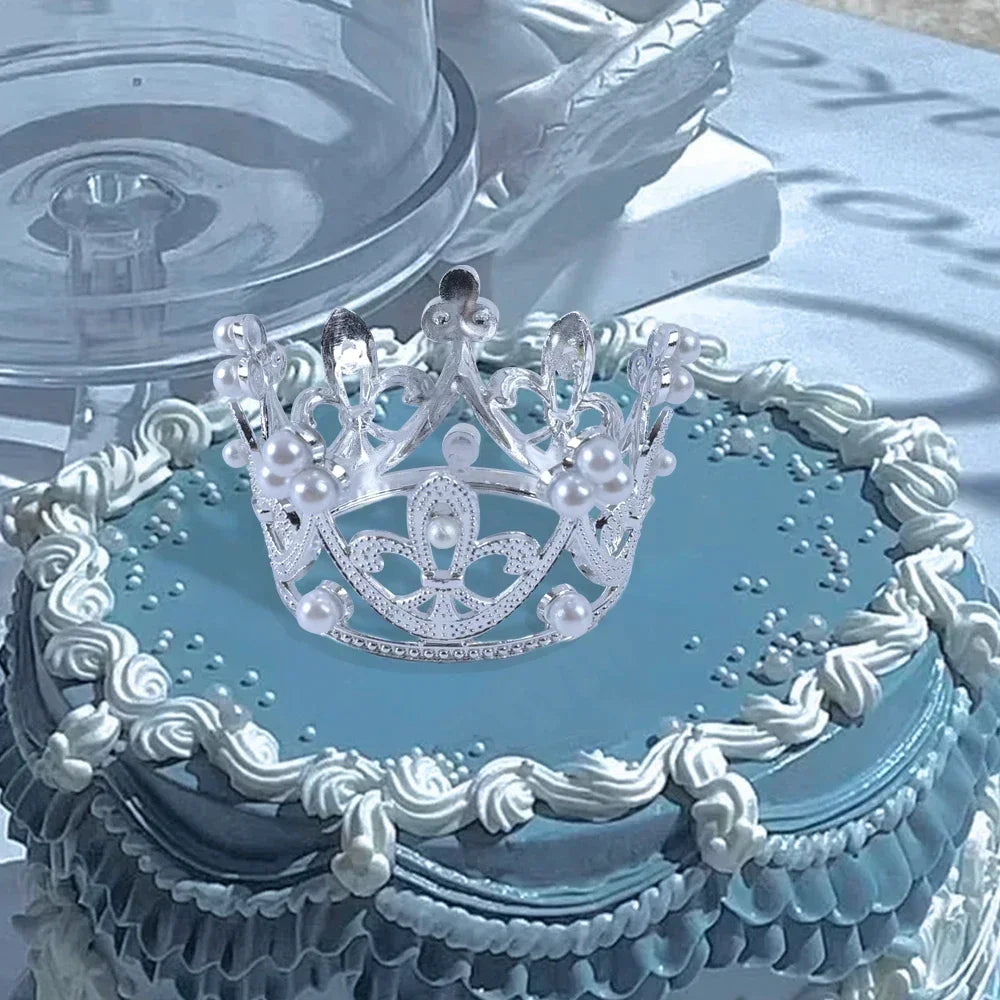 A tiered cake decorated with blue frosting and silver lace, topped by a sparkling mini gold and silver crown.