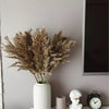 A dried pampas grass bouquet in a white vase sits on a light surface next to a candle and an alarm clock.