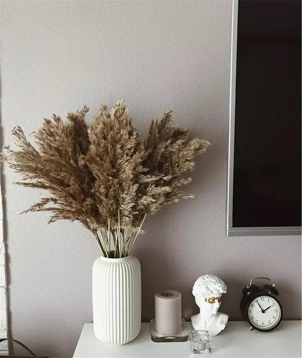 A dried pampas grass bouquet in a white vase sits on a light surface next to a candle and an alarm clock.