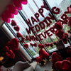 A display of red foil birthday balloons shaped like hearts with bows hanging in front of a window.