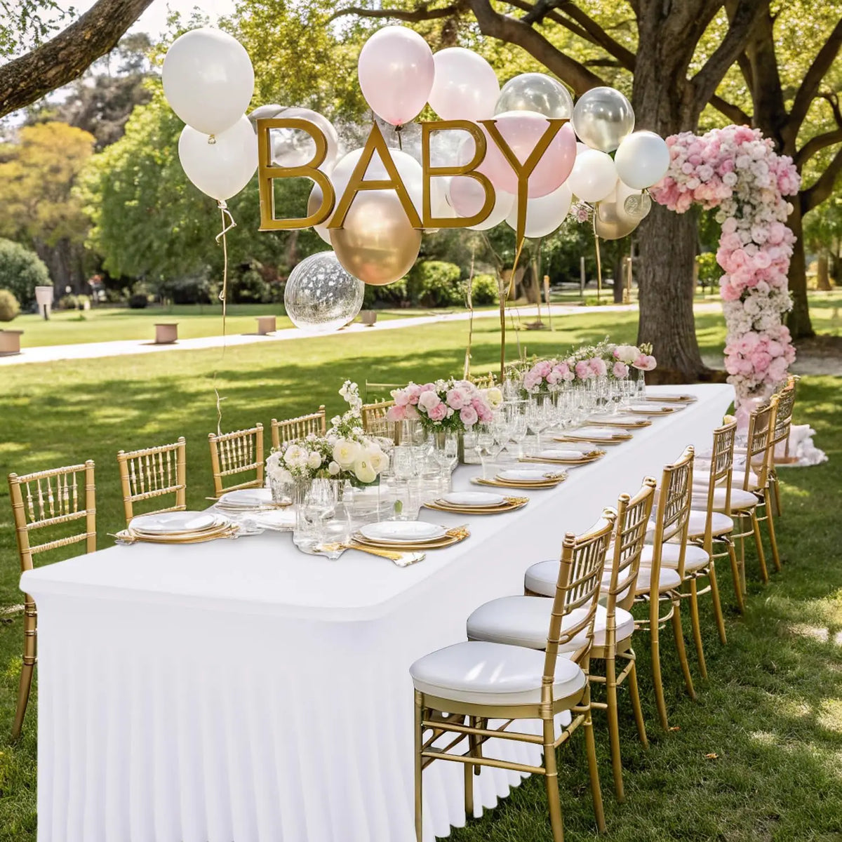 A long white table set for an outdoor event featuring gold chairs and balloons in a park setting.