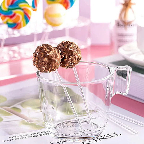 Clear acrylic cake pop sticks with chocolate truffles are displayed on a pink surface.