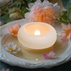 A white floating LED candle glows warmly on a ceramic dish surrounded by pink and white flowers.