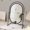 A black oval vanity mirror with a thin metal frame and a reflective surface is shown on a white table.