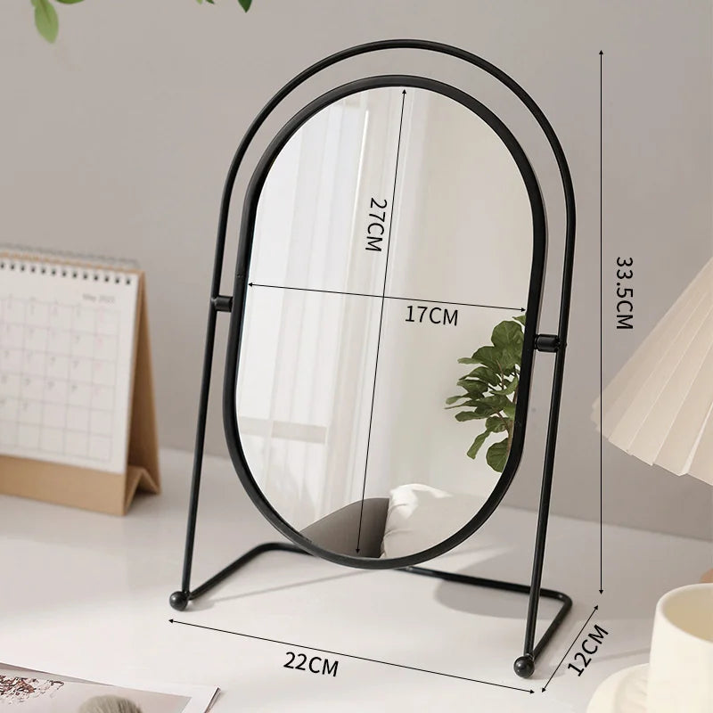 A black oval vanity mirror with a thin metal frame and a reflective surface is shown on a white table.