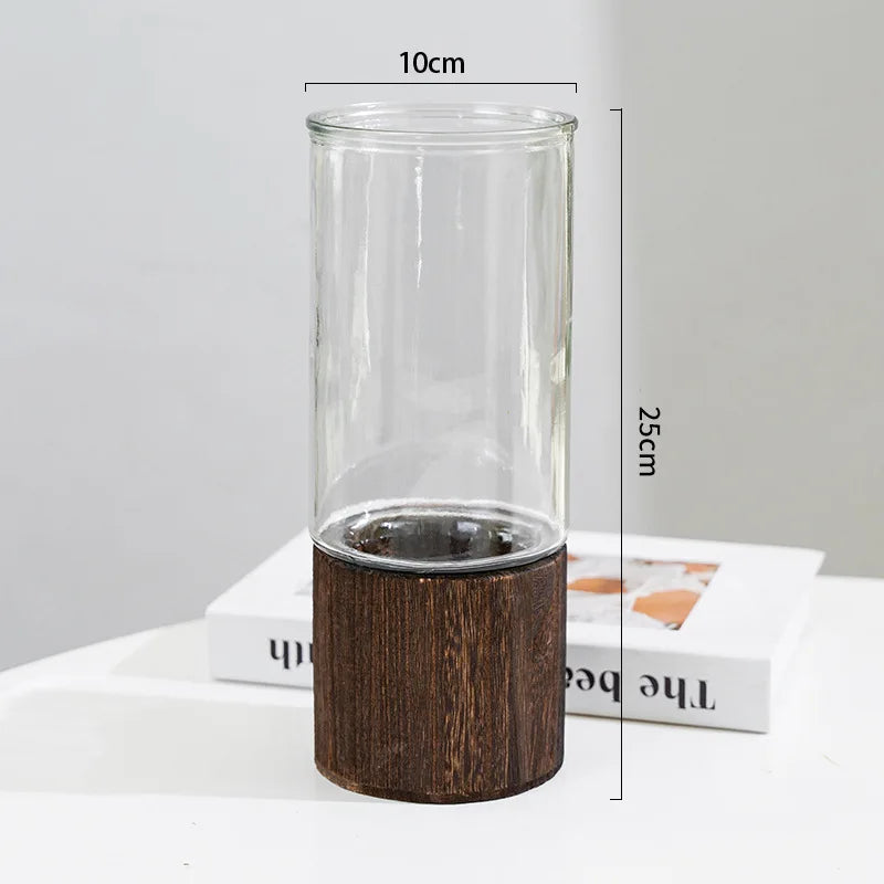 A tall, clear glass vase with a dark wooden base is shown standing upright on a white surface.