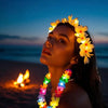 A woman wearing a glowing LED Hawaiian lei necklace with yellow flowers against a twilight sky and ocean backdrop.
