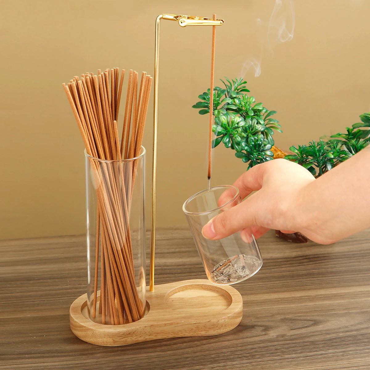 A hand pours incense sticks into a glass holder with a gold-colored metal rod and wooden base.