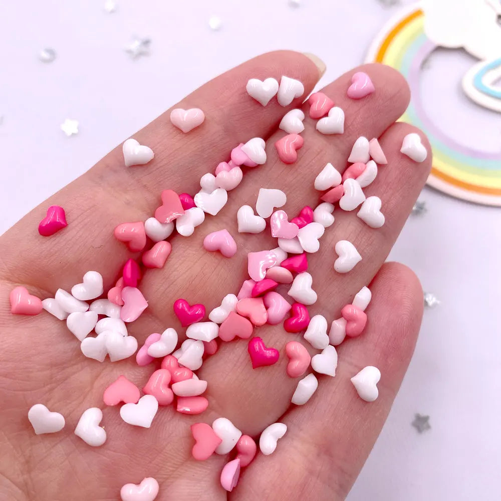 A hand holds a mix of pink and white heart-shaped flatback rhinestones for nail art and crafts.