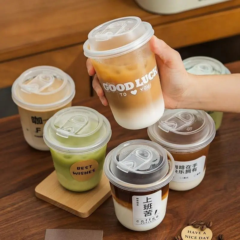 Una mano sostiene una taza desechable en forma de U con café helado, tapa y pajita, sobre una superficie de madera.