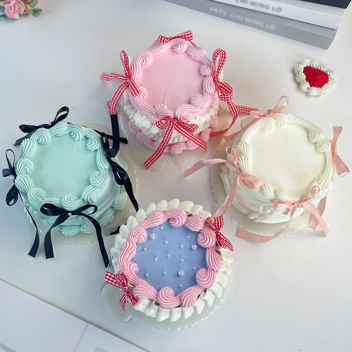 Four decorative fake cakes in pink, mint green, and white with floral piping and ribbons on a white surface.