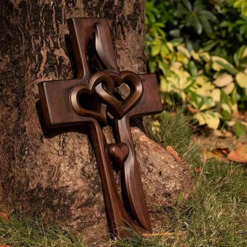 A dark brown wooden wall cross with intertwined hearts carved into the design, set against a tree trunk.