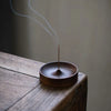 A thin copper incense stick is burning inside a dark wooden bowl on a wooden surface.