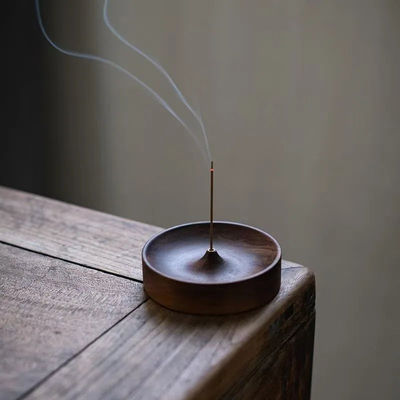 A thin copper incense stick is burning inside a dark wooden bowl on a wooden surface.