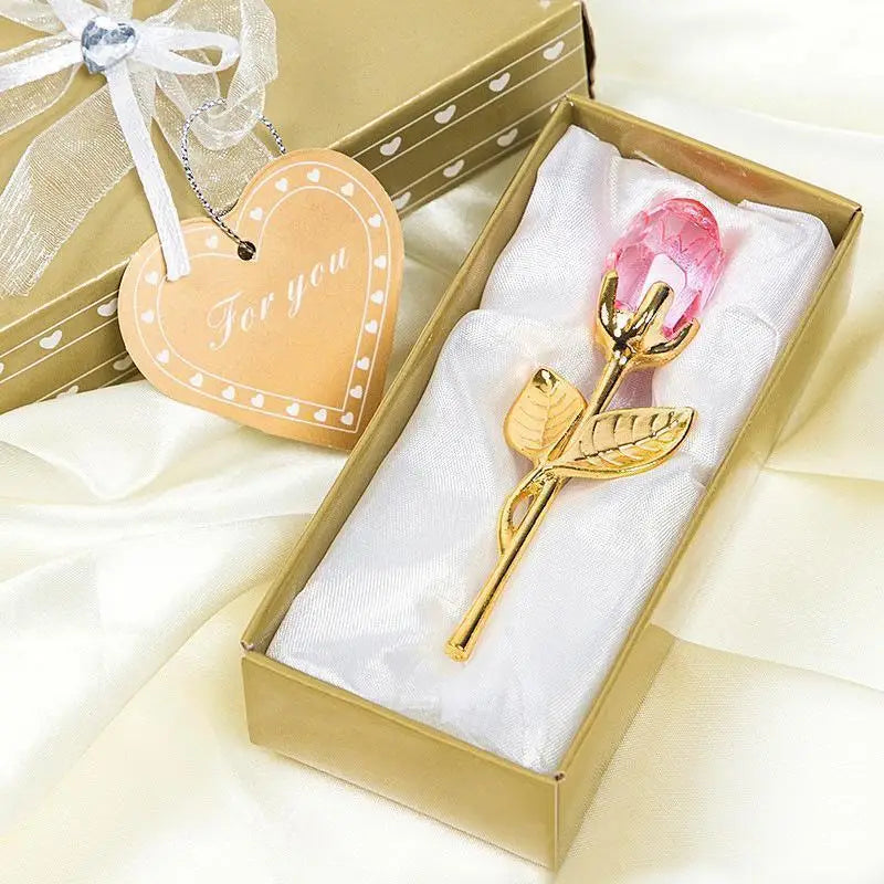 A gold box contains a crystal rose artificial flower in a white velvet insert and a heart-shaped tag.