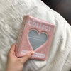 A pink multi-size photo album for collectibles with a heart-shaped clear window is held in a hand on white bedding.