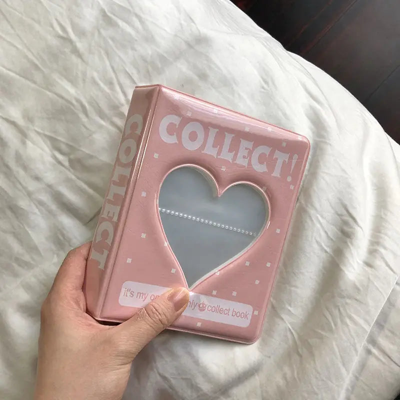 A pink multi-size photo album for collectibles with a heart-shaped clear window is held in a hand on white bedding.