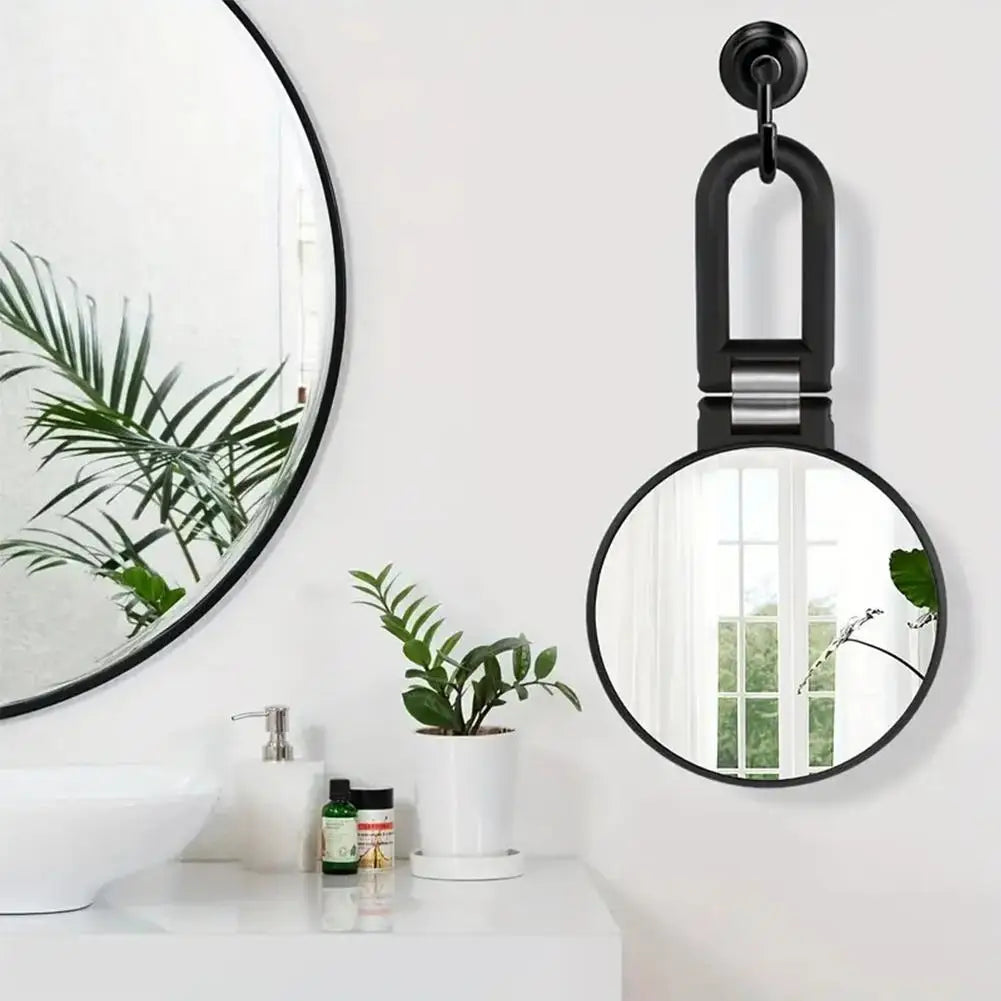 A black precision dual-magnification handheld mirror hangs on a white wall above a bathroom vanity.