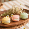 A wooden platter displays several macaron-shaped soy wax candles in pastel pink and green colors with dried flowers.