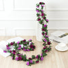 A long garland of deep purple silk roses draped vertically against a white vase on a light wooden surface.
