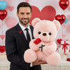A man holds a pink rose bear plush toy with red ribbon and faux flowers against a pink heart background.