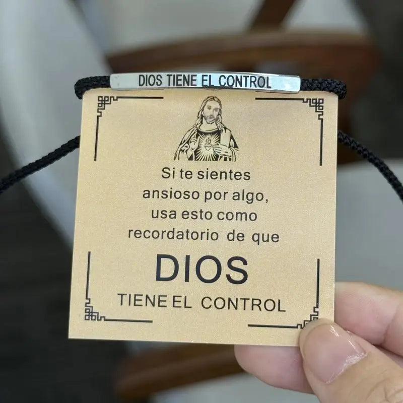 A beige leather tag with the text Dios Tiene El Control is attached to a black cord, used as an inspirational bracelet.