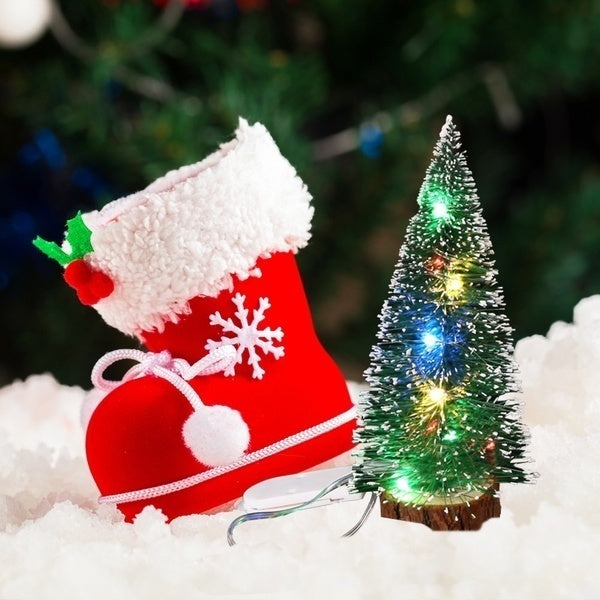 A red Christmas stocking with white fur trim and a green LED mini tree decorated with lights in a snowy setting.