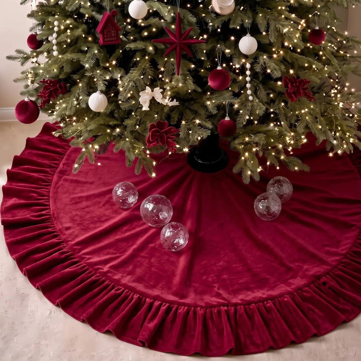 A velvet red Christmas tree skirt with a ruffled edge surrounding a decorated green fir tree.