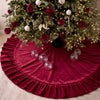 A velvet red Christmas tree skirt with a ruffled edge surrounding a decorated green fir tree.