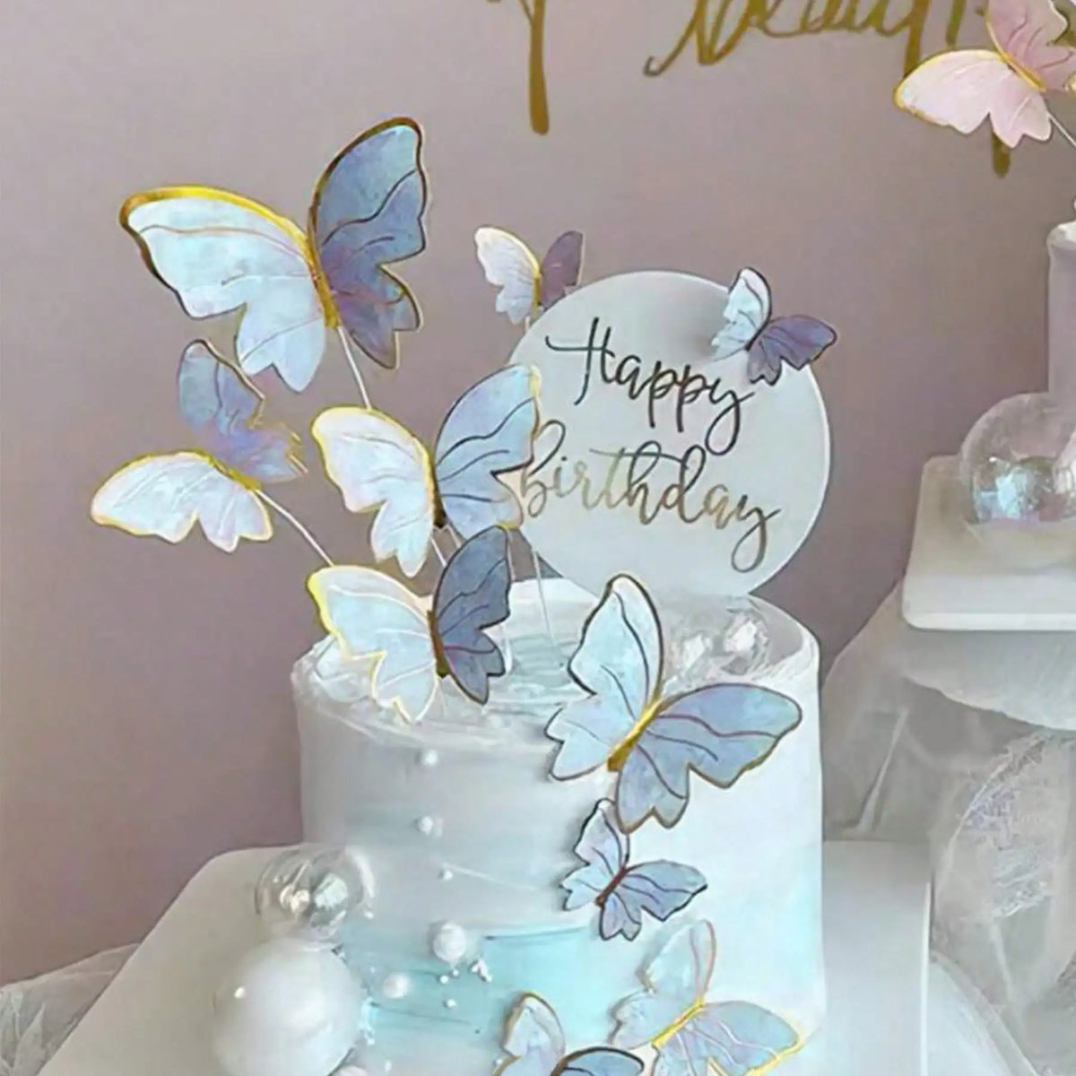 A white cake decorated with blue and gold butterfly toppers on a light pink background.