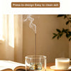 A glass incense burner with a long metal stick is shown emitting smoke on a wooden table.