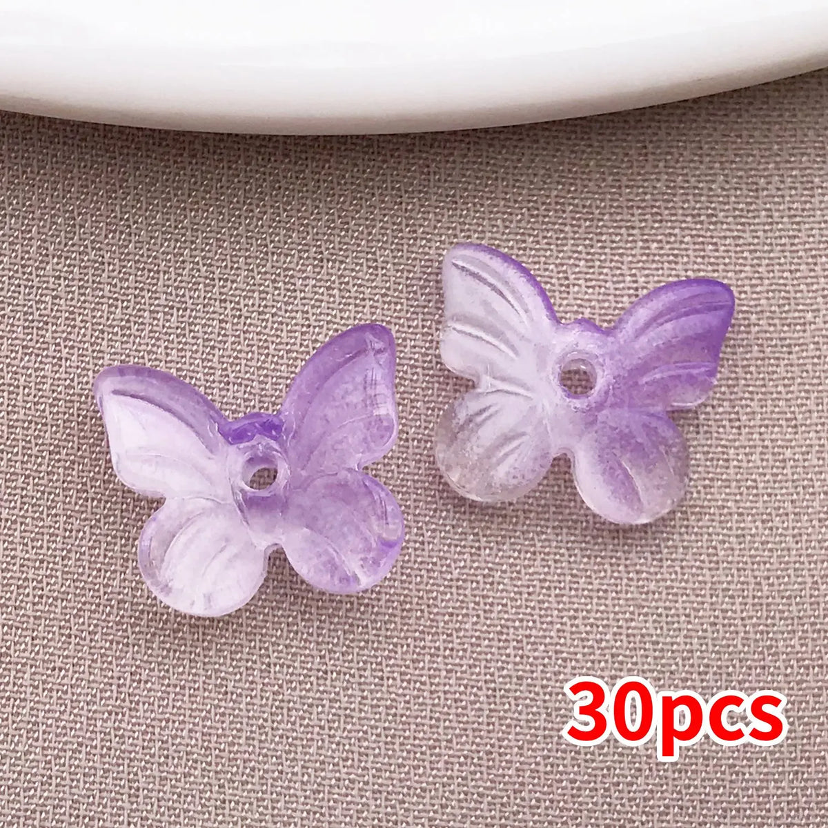 Un juego de 30 cuentas de mariposa de vidrio morado sobre una superficie de tela gris.