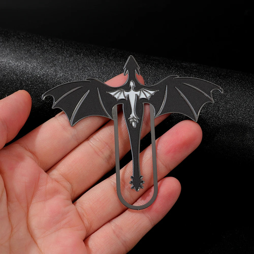 A black metal bookmark shaped like a four-winged dragon is held in a person's hand against a dark background.