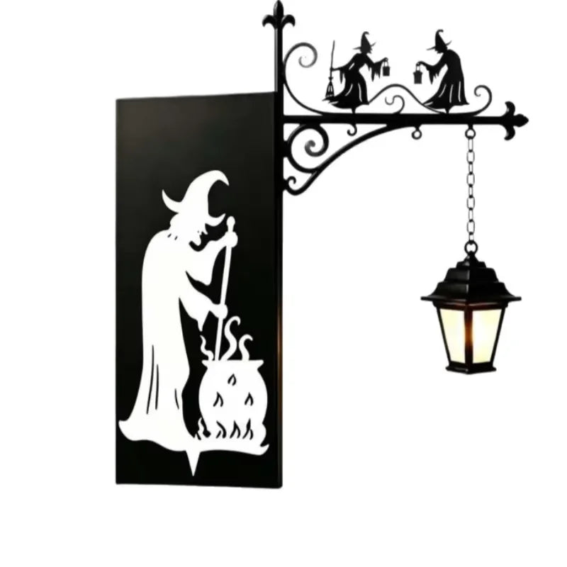 A black silhouette of a witch and cauldron design mounted on a wrought iron bracket with a hanging lantern light.