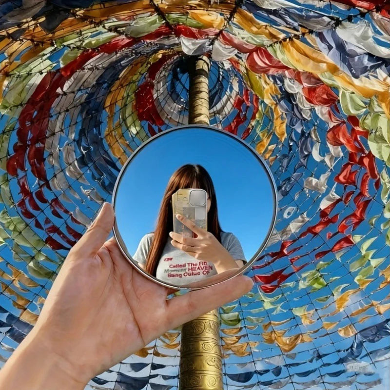 A hand holds a round convex mirror reflecting the sky and colorful fabric above.