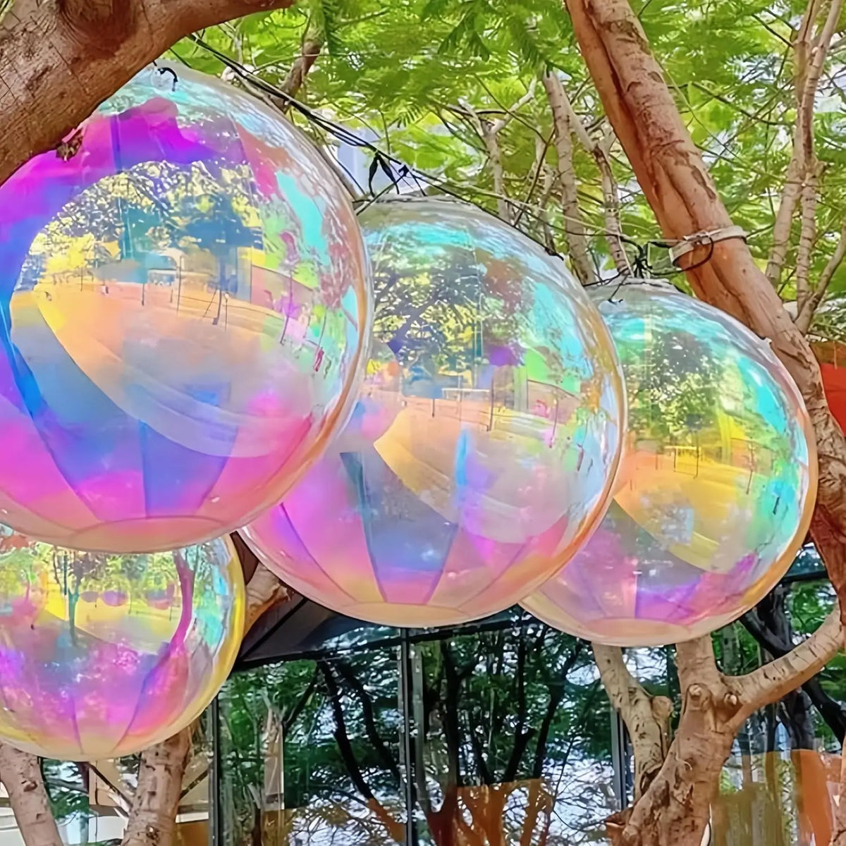 A cluster of iridescent holographic round foil balloons hanging from tree branches in an outdoor setting.