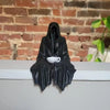 Black resin statue of a ghost figure holding a white teacup against a brick background.