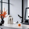 Statue fantôme blanche décorative sur table blanche, à côté d'une citrouille orange et d'une figurine de chauve-souris noire.