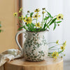 A rustic ceramic milk pitcher with a cracked glaze and pastoral design filled with yellow flowers.