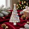 A sparkling rhinestone Christmas tree brooch pin displayed on a red velvet fabric with pine branches and gold ornaments.
