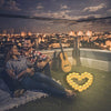A couple sits on a rooftop overlooking a city at night with a heart shape made of glowing tea light candles.