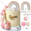 A white cake decorated with pink ball and cloud toppers, featuring a gold script name.