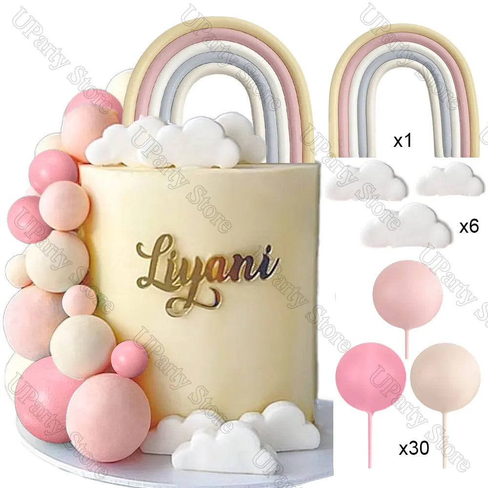 A white cake decorated with pink ball and cloud toppers, featuring a gold script name.