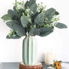 A bouquet of artificial eucalyptus stems with small white flowers displayed on a light green vase.