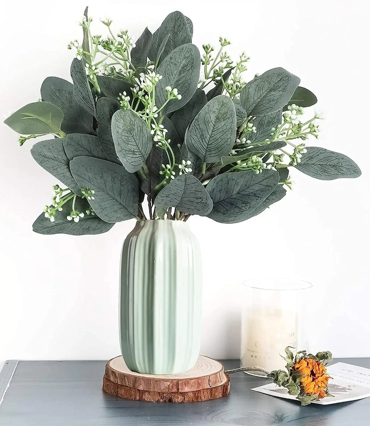 A bouquet of artificial eucalyptus stems with small white flowers displayed on a light green vase.