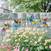 Colorful metal hummingbird wall art displayed against a stone wall with a garden of white and yellow flowers.