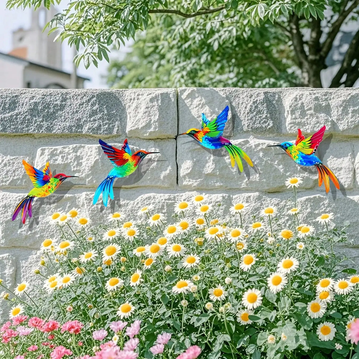 Colorful metal hummingbird wall art displayed against a stone wall with a garden of white and yellow flowers.