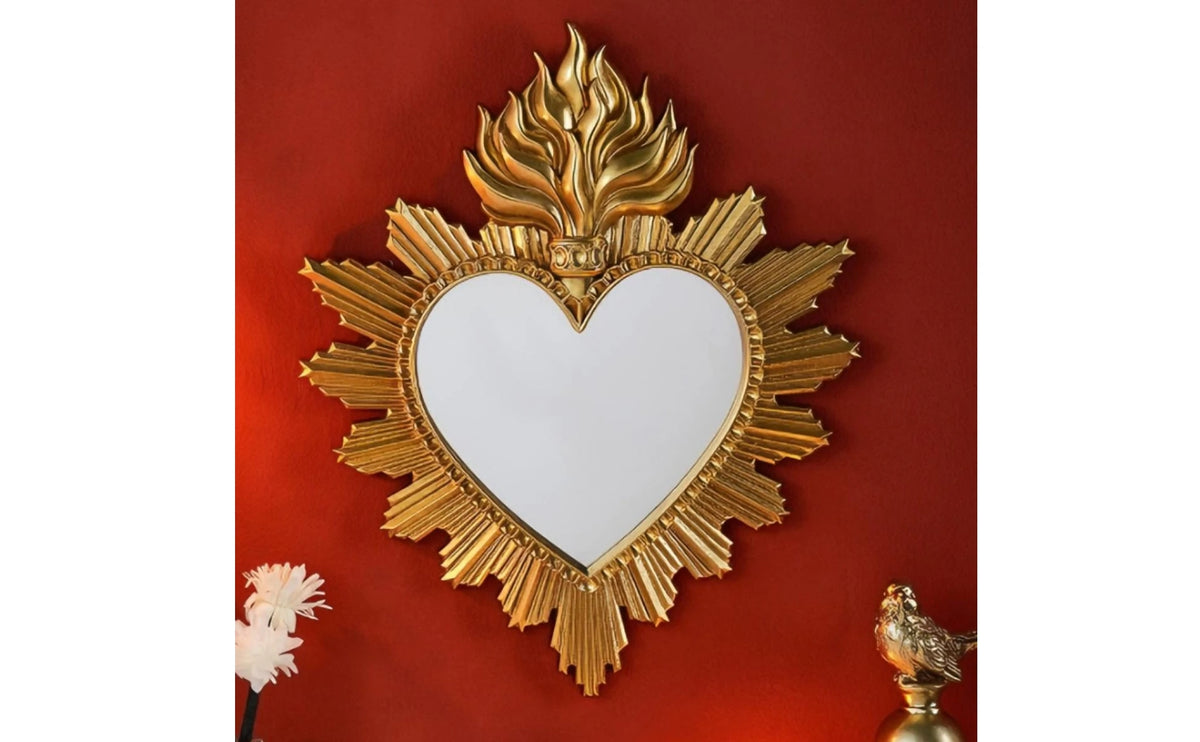 Ornate gold-framed heart-shaped wall mirror with flame detailing mounted on a red wall.